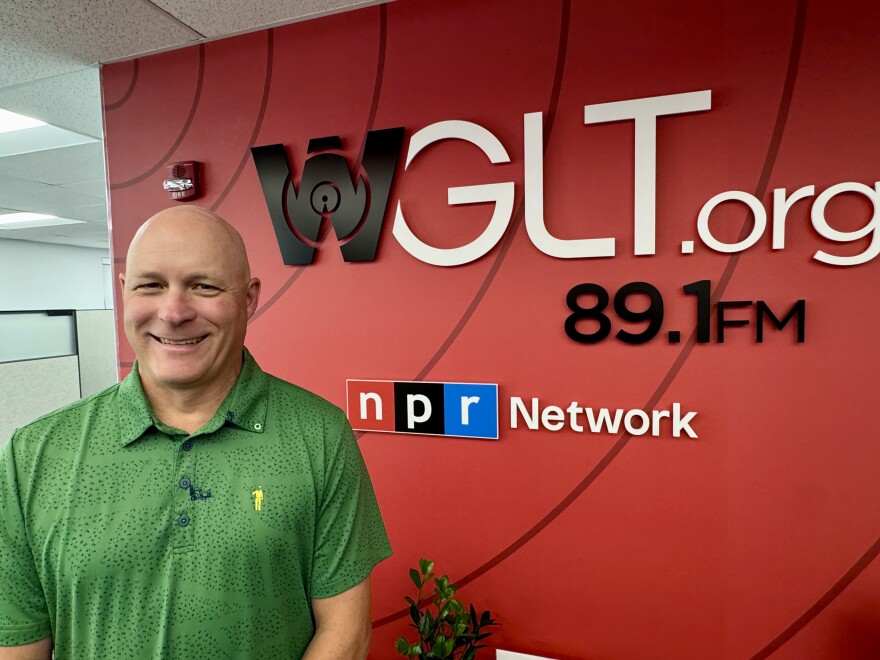 A smiling man in a green shirt stands in front of a red wall with the WGLT.org 89.1 FM and NPR Network logos. Office cubicles are visible in the background.