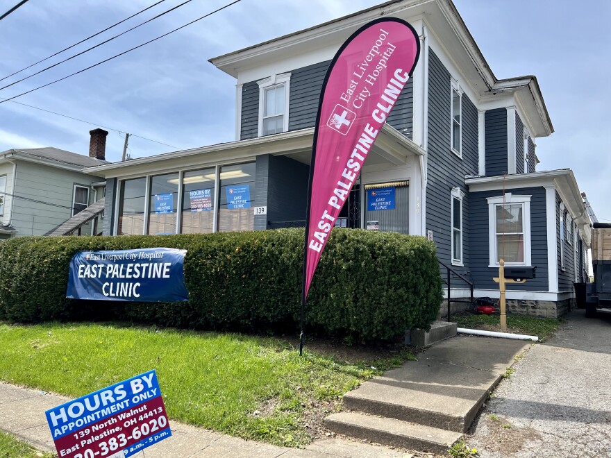 A blue building with green bushes in front advertises the "East Palestine Clinic". A sign in the yard details the hours and number to call for appointment.