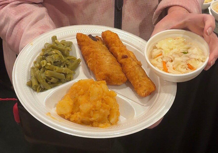 A customer at a local fish fry shows off her fried cod, green beans, mashed potatoes, and coleslaw on a styrofoam plate.