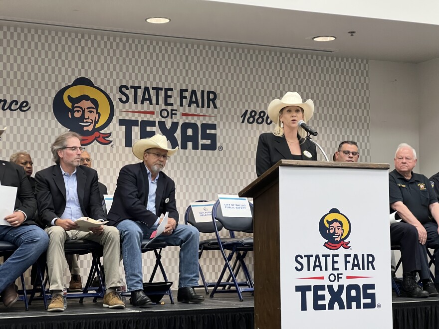 A woman in a cowboy hat stands behind a wooden podium, which has the words "State Fair of Texas" and a picture of a man in a cowboy hat and scarf on the front. Behind the woman are several people sitting in chairs on a stage.