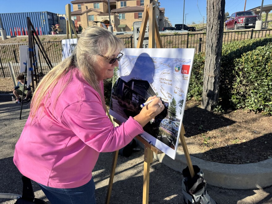 A woman wearing sunglasses signs her name to an artist's rendering of a modern building. The rendering is on a poster and easel.