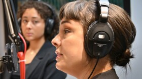 Two women sit inside a radio studio at microphones. The one on the right is talking while the other looks at her and listens.