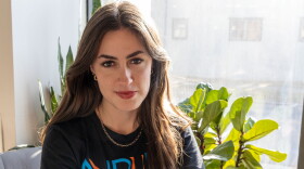 A young woman with long, brown hair gives the camera a half-smile. She's wearing an NPR t-shirt, a gold chain necklace and gold hoop earrings. Behind her are large-leafed plants and a sunlit (kind of dirt-streaked, if we're being honest) window.