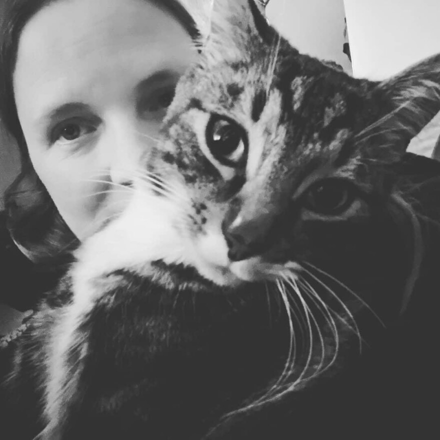 Μια ασπρόμαυρη φωτογραφία μιας γάτας που κοιτάζει απευθείας στην κάμερα με ένα άτομο πίσω από τη γάτα.