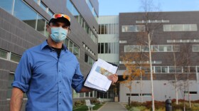 A man in a blue button up shirt, black baseball cap, and sunglasses, holds a piece of paper with photos of an apartment.