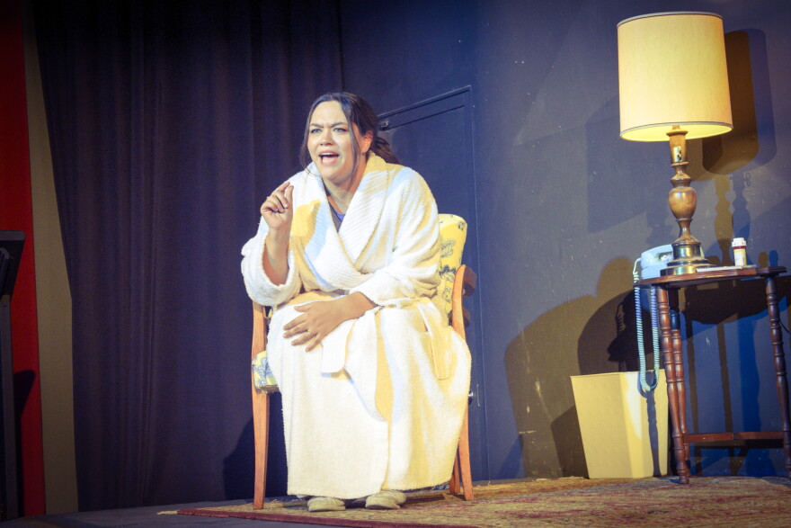 A woman in a white bathrobe sits on a chair, gesturing with her finger as if speaking passionately. She is barefoot, and a lit lamp and small table are beside her. The scene appears to be from a stage play.