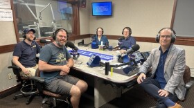 Five smiling people wearing headphones sit at a table in a radio talk studio: a man front left has a light brown beard and mustache and is wearing a blue baseball hat, a blue polo shirt, and khakis; a man front center has short dark hair and a black and grey mustache and beard and is wearing glasses, a t-shirt with an image of a bike on it; grey shorts, and black sneakers; a woman back left has short brown hair and is wearing a blue top; a man back center has short brown hair and is wearing glasses and a blue button-down shirt; a man at right as very short dark hair and is wearing a blue button-down shirt under a grey plaid suitcoat, jeans, blue socks, and brown shoes