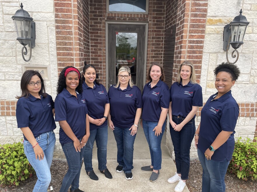 A group of women stand in front of an office in jeans and blue shirts.