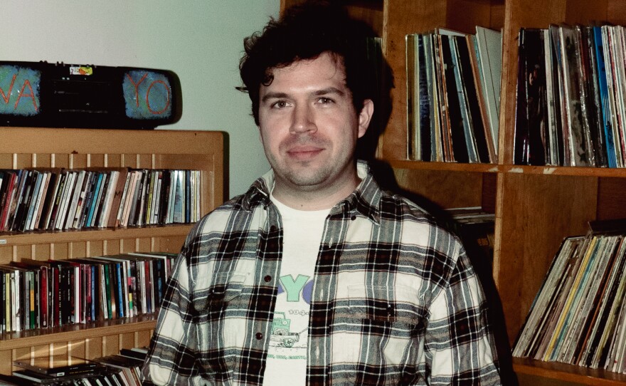 A man with brown hair wearing a graphic t-shirt under a white and black flannel shirt. He is standing in front of bookcases of CDs and LPs.