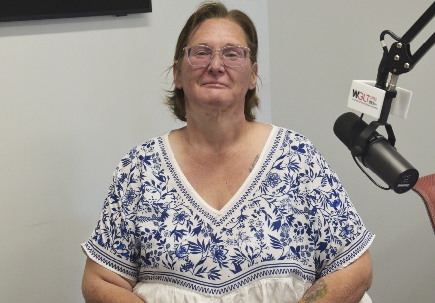 Woman in short hair and glasses sitting next to a microphone and wearing a v-neck white blouse with blue flower patterns 