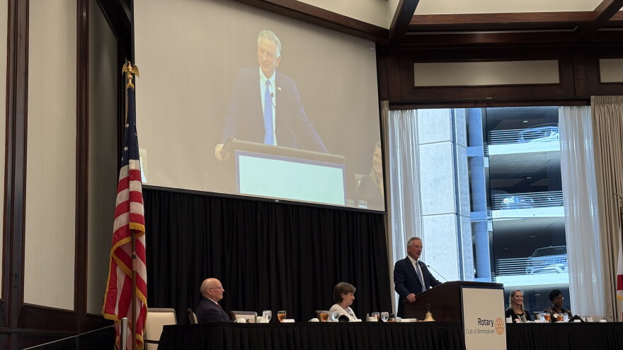 Tommy Tuberville stands behind a podium with people along a table on both sides of the podium. A screen showing Tuberville is above him.