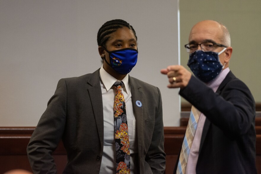 A woman and a man in masks and suits stand next to each other. The man, who is on the right, is pointing at something out of view.