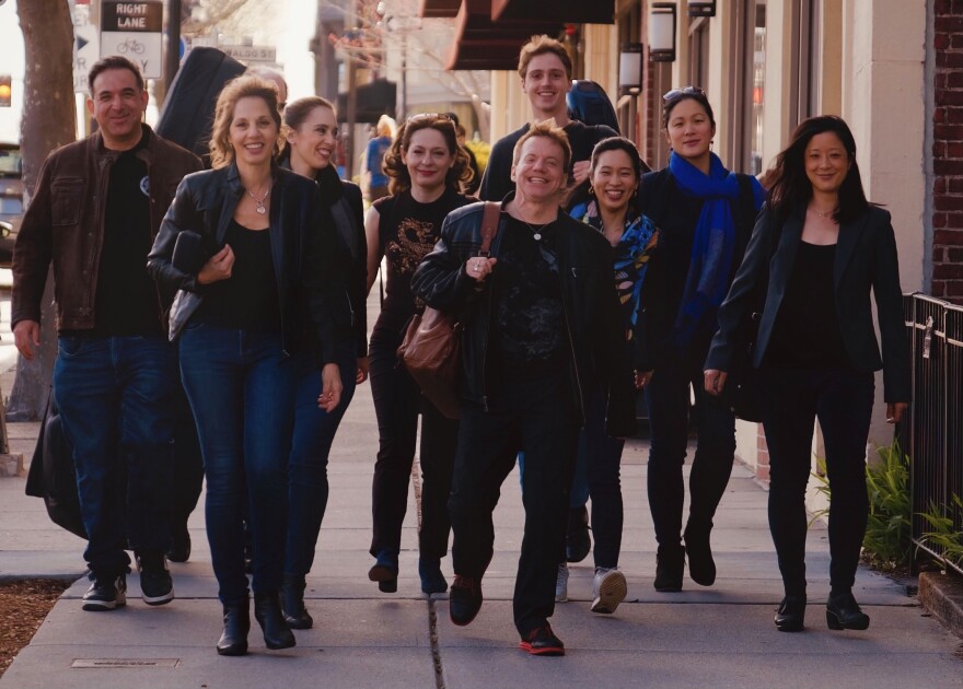 Members of Mistral walk down a sunny sidewalk with their instruments. They wear black and blue, and they smile at each other and the at the camera.