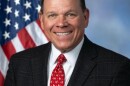A man, Rep. Sam Graves of Missouri's 6th District, is dressed in a black suit and red tie, smiling, seated for a portrait in front of a draped American flag backdrop.