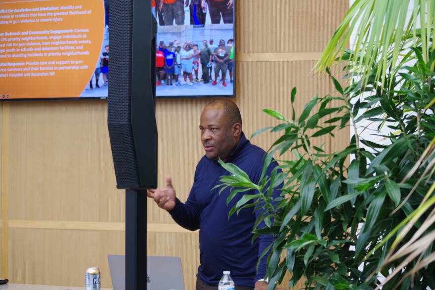 Reggie Moore, Director of Violence Prevention Policy and Engagement for the Medical College of Wisconsin Comprehensive Injury Center wears a blue violence prevention shirt.  He's at the Winter Garden in the Kalamazoo Community Foundation's office.  Standing near plants and in front of his presentation screen.  A black speaker can be seen in the shot. 