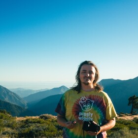 Smiling man standing in a tye-die shirt in front of a mountain range in the sun 