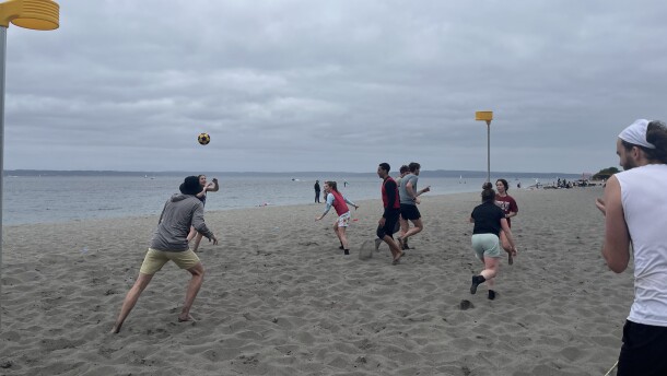 People throw a ball up and run around on a beach on a cloudy day.