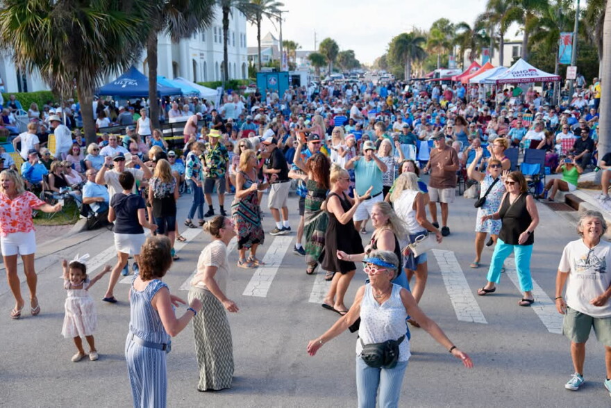 A large crowd fills a palm tree-lined street at an outdoor evening concert, with dozens of people dancing in the foreground and spectators seated in lawn chairs along the sidewalks under vendor tents in the background.