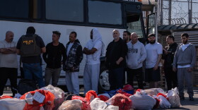 A number or men line up next to a bus while a man with a 'Police ICE' vest speaks to some of them. 