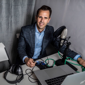 A man with brown hair wears a blue suit and sits at a table with a microphone, computer, and headphones on it. 