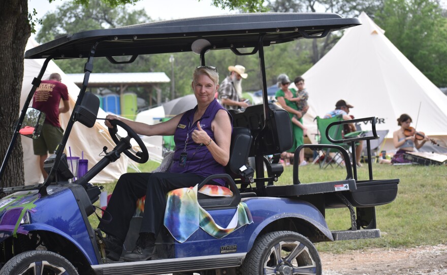 Kerrville Folk Festival Executive Director Deb Rouse rides through Quiet Valley Ranch in a golf cart as musicians warm up behind her.
