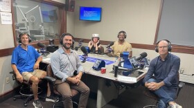 Five people wearing headphones sit at a table in a radio talk studio: a man front left has short brown hair and is wearing a bright blue polo shirt, khaki shorts and sneakers; a man front right has short dark hair, a dark beard and is wearing a light blue button-down shirt, brown pants and brown shoes; a woman back left is wearing a white hat, eyeglasses and a black sleeveless top; a man back right has very short dark hair and is wearing a beige polo shirt with white trim; a balding man at center is wearing eyeglasses, a blue button-down shirt, blue jeans and sneakers.