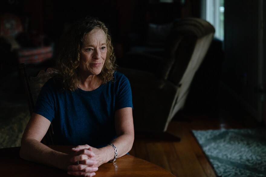 A woman wearing a blue T-shirt sits at a wood table with her hands clasped in front of her.