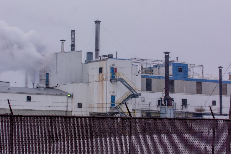 Industrial facility with white and blue exterior, multiple smokestacks emitting smoke, yellow staircases, and barbed wire fencing in the foreground on a cloudy day.