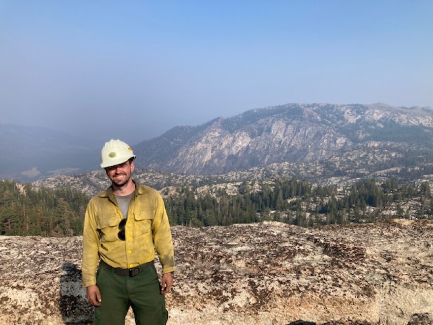 Snake River Hotshots third-year crew member Brigham Snow poses smiling wearing a white hard hat with forested mountainous terrain in the background.