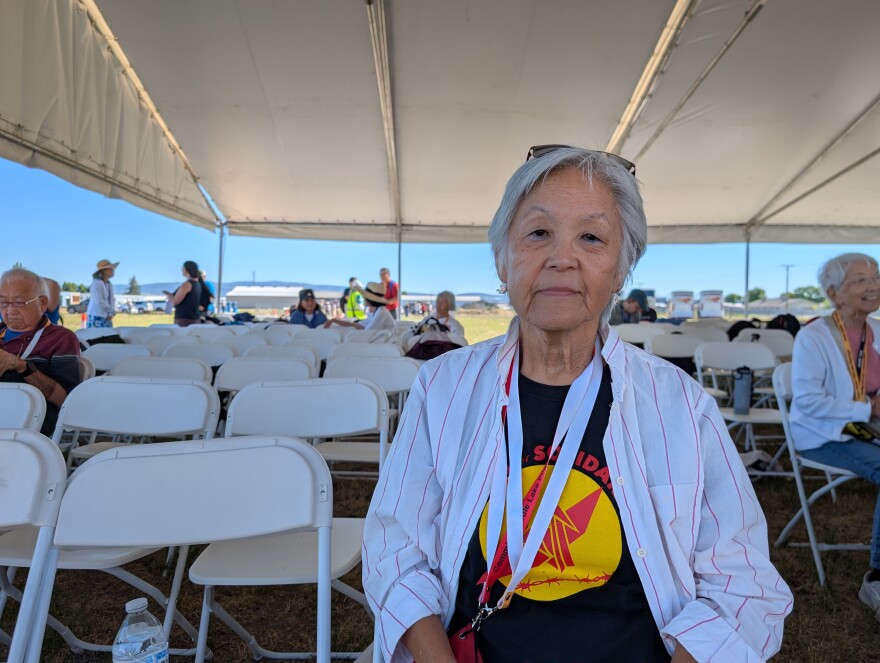 A woman wit grey hair sits on a folding chair with others in the background