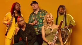 Five musicians pose for the camera, wearing stylish clothing and holding their instruments, before a gold background