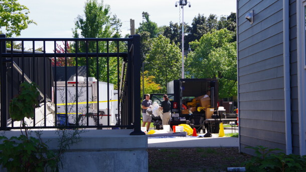 Two individuals can be seen holding a large, plastic object near several trailers in a building parking lot.