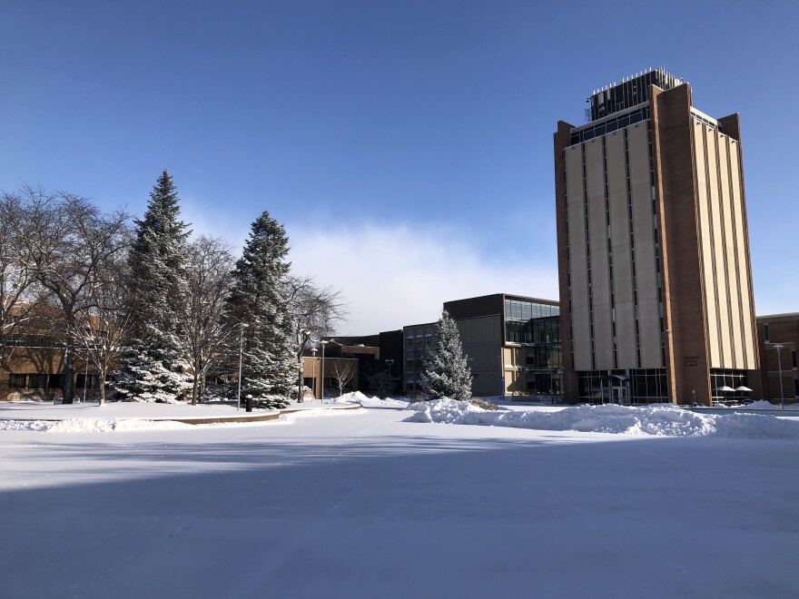 Picture of open, plaza-type space with a light coating of snow, clear sky, and buildings in the background including one on the right that is quite tall.