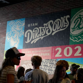 A colorful mural on a brick wall reads "Utah Dirty Sodas" and "2026." A crowd of people stand in front of it, facing different directions. 
