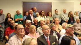 A crowd of people, most of them middle aged, sit and watch a town council meeting. 