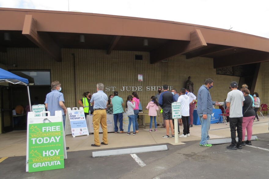 Community members stand in the parking lot in front of a church. They are standing in line and filling out forms on clipboards. 