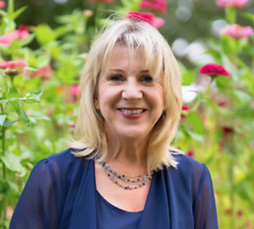 Angela Corell with long blonde hair wearing a blue top. Backdrop is a flower garden.