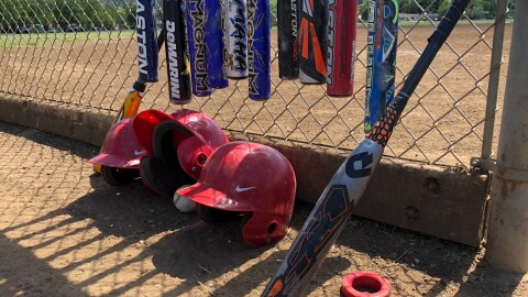 Baseball equipment such as helmets and bats lean up against a chain link fence.