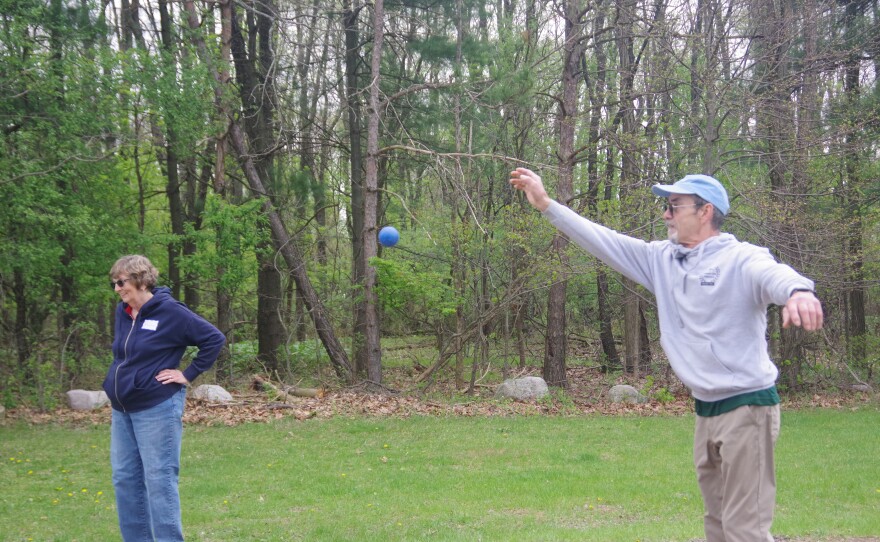 Man holds arm up in the air as he lets go of a blue boule which is in the air in front of him, while a woman stands nearby in blue jeans and dark blue jacket