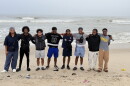 A group of young men with their arms around each other, standing on sand in front of a body of water