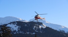 A red-and-white helicopter with a ski basket descending to land with snowy mountains in the background