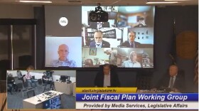 A screenshot of lawmakers faces projected over a screen, as they call into a Zoom meeting, in a legislative meeting room.