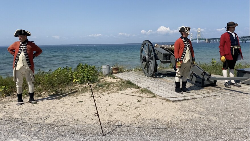 A man in a British uniform stands to the left of two men, also wearing historical garb. The two men are in front of a cannon pointed towards Lake Michigan. The Mackinac bridge is in the background.