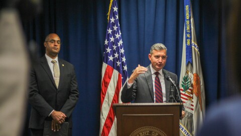A man in a suit and tie speaks at a podium, with a U.S. flag and another flag behind him. The second flag has a banner that reads "Southern District of Ohio." A second man in a suit and tie stands to the side, hands folded. 