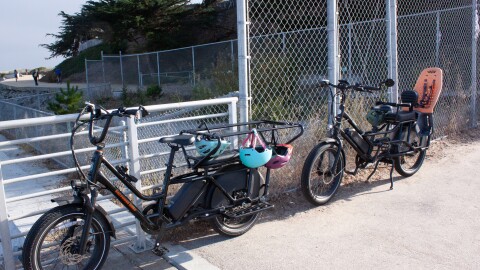 Two large e-bikes are parked against a fence with helmets hanging from them. One has a long seat for two kids and the other has a seat with a harness for one child. 