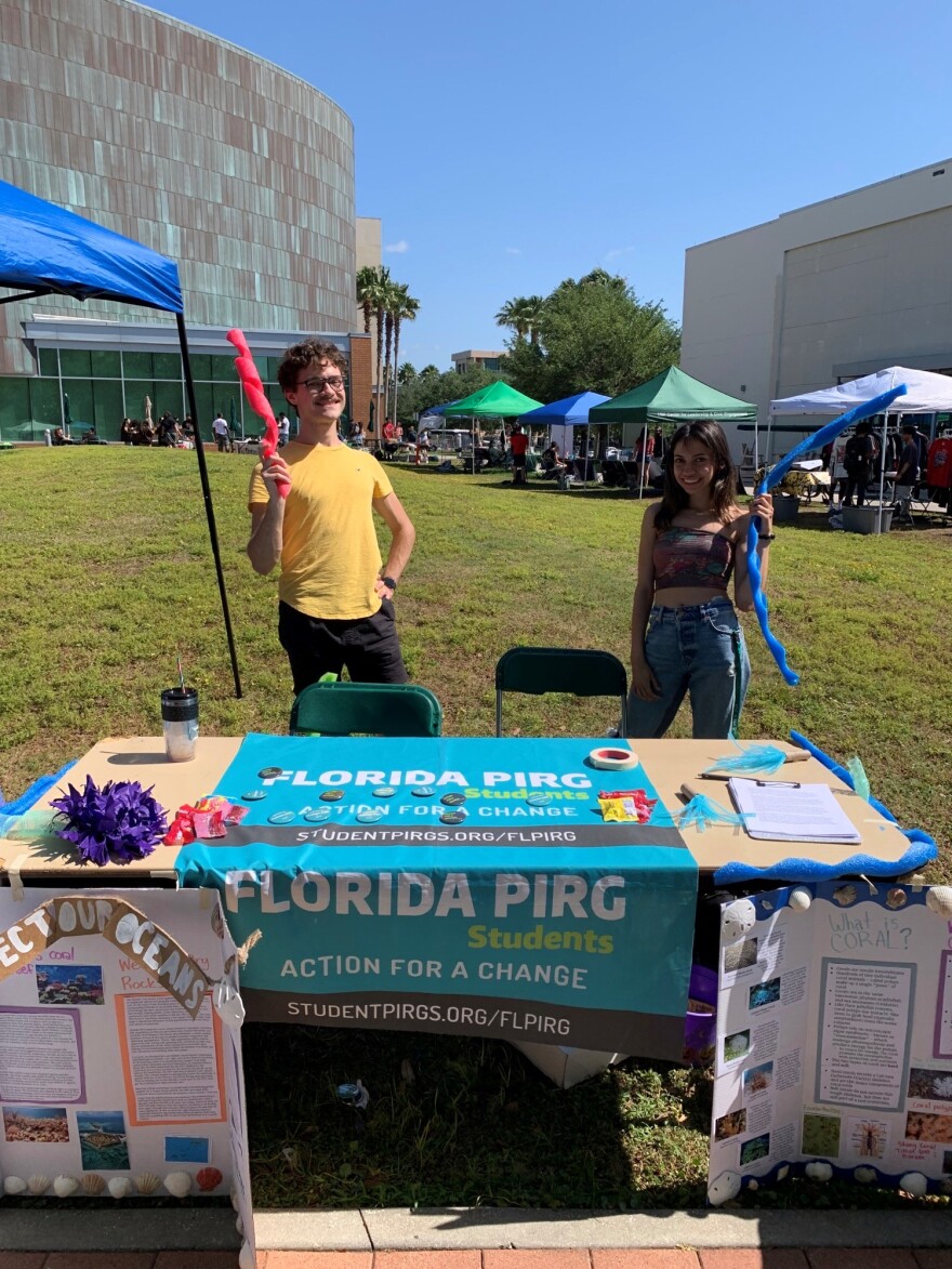 On a sunny day with clear skies, there are tents and tables set up across the green grass. At the center of the photo stand two students behind a table with information about Florida PIRG and renewable energy.