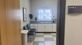The image shows a doorway leading into a brightly lit bathroom.  There is a white sink across the room directly opposite the doorway.  There is a small window  above the sink.  In the foreground, there are two metal folding chairs with blue upholstery.  There is text on the wall above the doorway that reads “EXAM 2”.