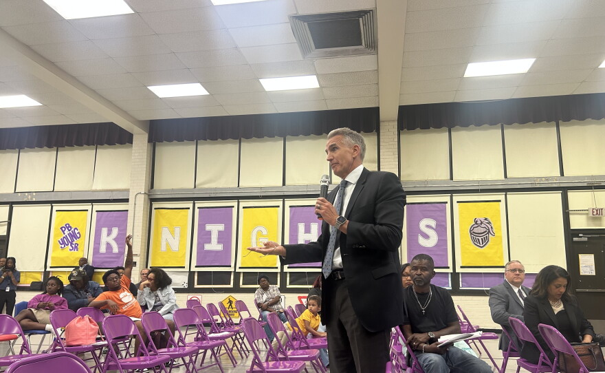 An image of Norfolk Public Schools Superintendent Jeff Rose standing in the cafetorium of P.B. Young Elementary holding a microphone while answering questions about a school consolidation plan. 