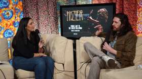  A woman and a man sit on separate couches while holding microphones during an interview.