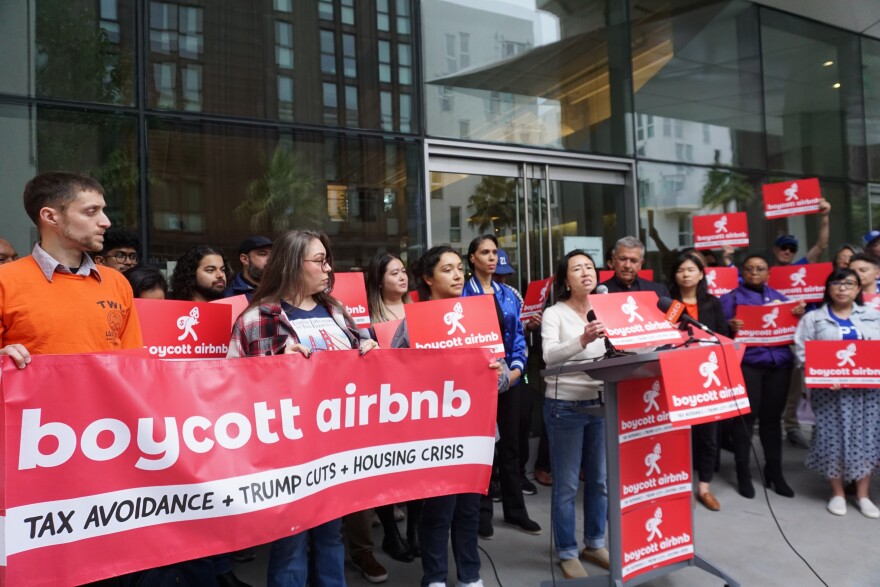 District 1 Supervisor Connie Chan speaks at a protest in front of Airbnb wearing a white shirt and jeans, surrounded by red and white protests signs that read "boycott airbnb."